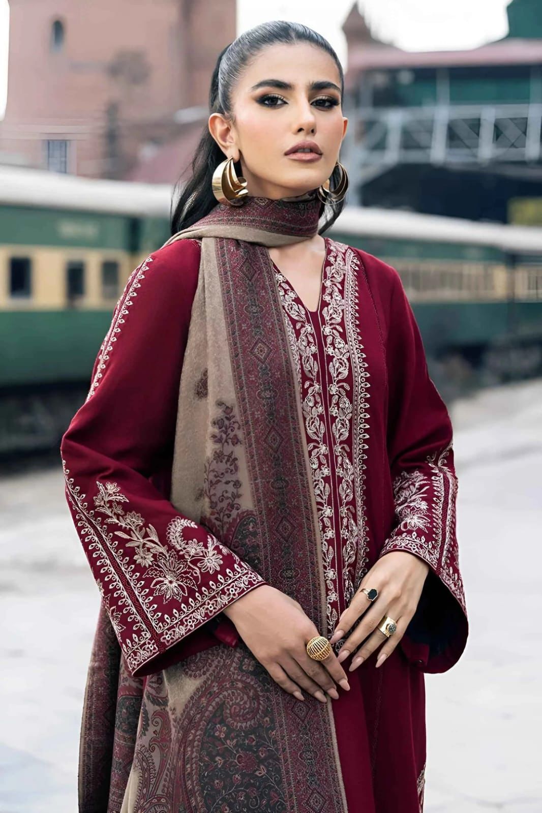 Woman wearing a maroon and brown traditional outfit with embroidery, standing in front of a train.